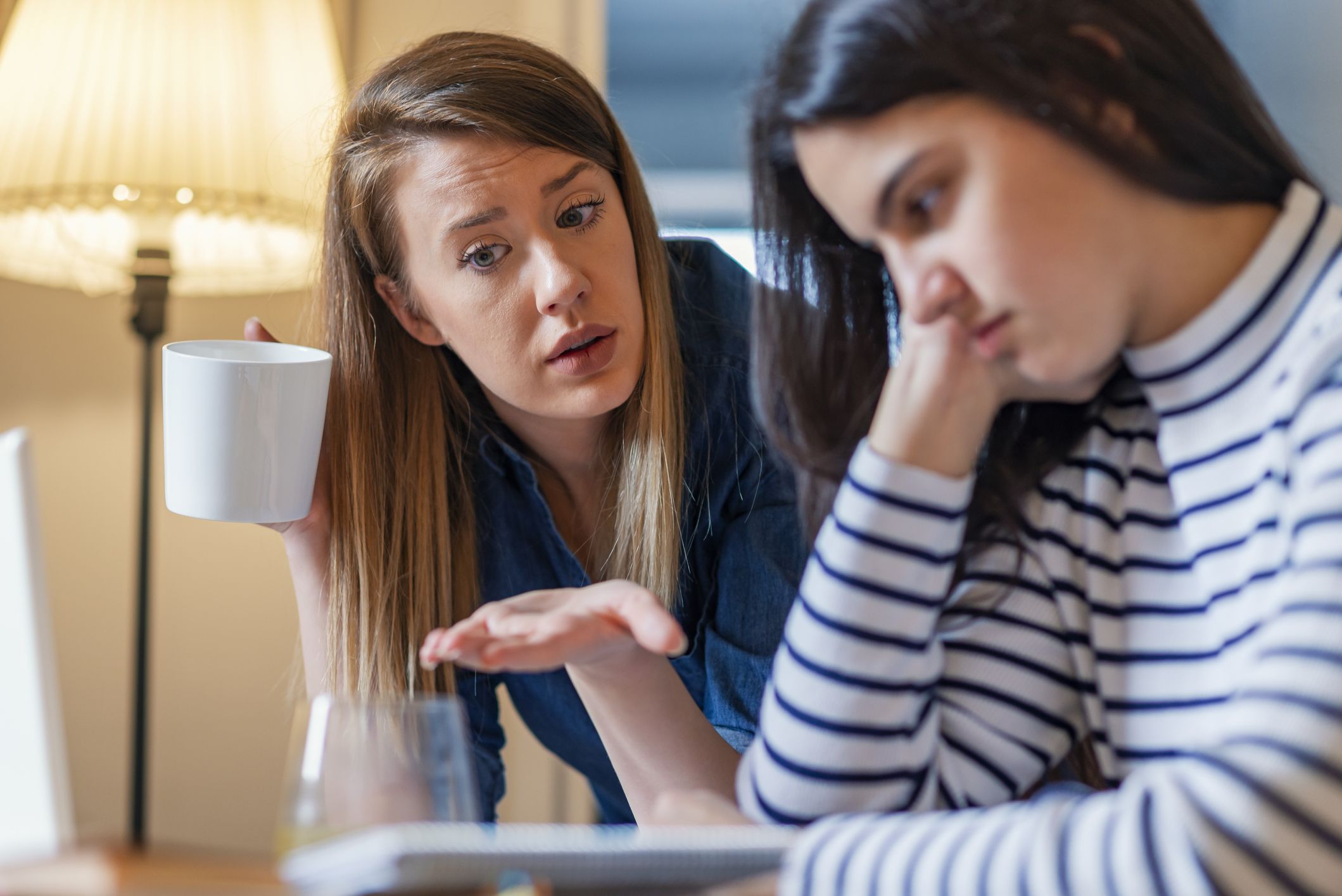Two women sitting at a table, one holding a mug while the other looks down.